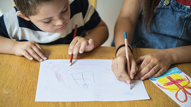 Un bambino con volto visibile e una bambina con le mani visibili, entrambi con matita in mano, appoggiati su un tavolo, che disegnano su un foglio
