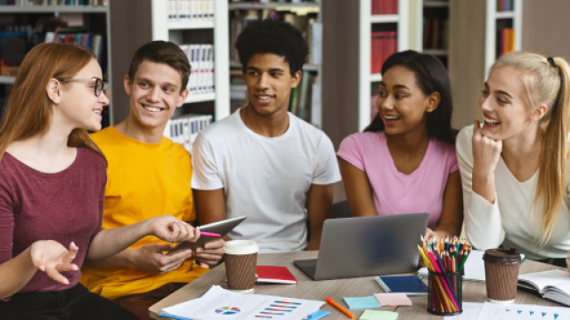 Gruppo di studenti che discutono seduti intorno ad un tavolo sul quale c'è un computer e un astuccio con delle matite. Uno degli studenti regge un tablet in mano