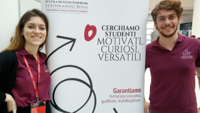 Una studentessa e uno studente a lato di un roll up che promuove la scuola di studi superiori