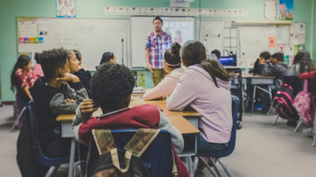 Svolgimento di una lezione in una classe media. L'insegnante è in piedi che spiega e alunni e alunne sono seduti e ascoltano