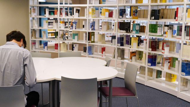 Studente intento a leggere un libro su un tavolo di una biblioteca. Di fianco a lui uno scaffale con libri.