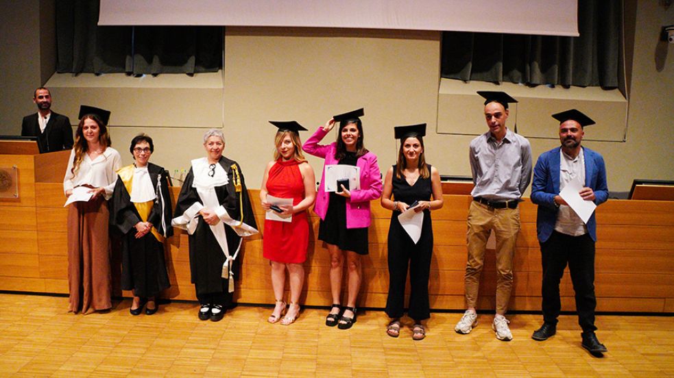 Sei persone con indosso il cappello da laureato, la prorettrice e una professoressa in toga posano di fronte al fotografo nell'aula magna della Cavallerizza in occasione della Festa della Conoscenza - Migliori Laureate e Laureati