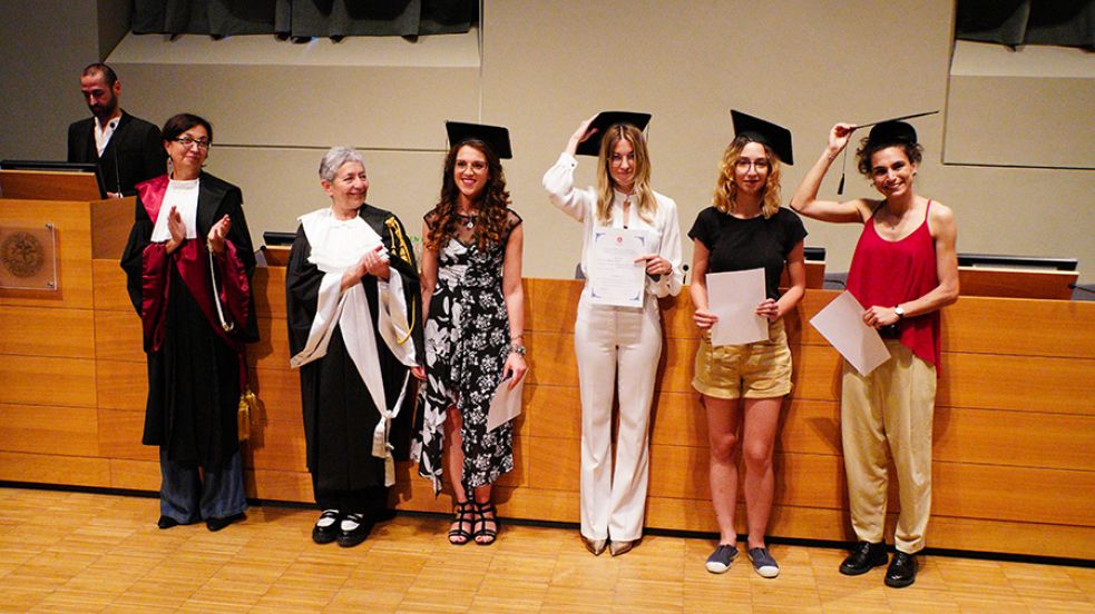 Quattro persone con indosso il cappello da laureato, la prorettrice e una professoressa in toga posano di fronte al fotografo nell'aula magna della Cavallerizza in occasione della Festa della Conoscenza - Migliori Laureate e Laureati