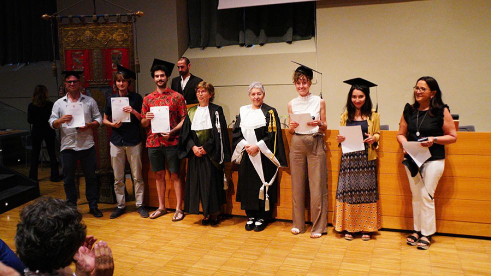 Sei persone con indosso il cappello da laureato, la prorettrice e una professoressa in toga posano di fronte al fotografo nell'aula magna della Cavallerizza in occasione della Festa della Conoscenza - Migliori Laureate e Laureati