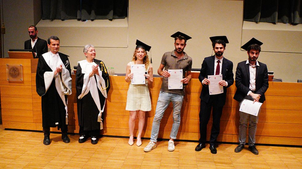 Quattro persone con indosso il cappello da laureato, la prorettrice e un professore in toga posano di fronte al fotografo nell'aula magna della Cavallerizza in occasione della Festa della Conoscenza - Migliori Laureate e Laureati