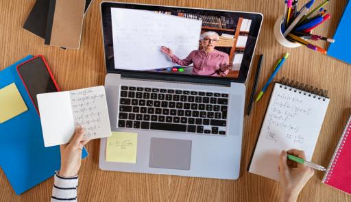 Immagine con computer portatile sul tavolo accesso con videolezione a distanza. Intorno al computer sono poggiati telefono cellulare, block notes, libri e portabiro. Si intravvedono le mani di una studentessa che reggono sulla mano sinistra un block notes con appunti e sulla mano destra una biro che prende appunti su un altro block notes