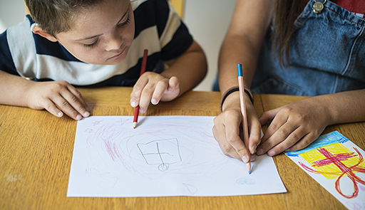 Un bambino con volto visibile e una bambina con le mani visibili, entrambi con matita in mano,  appoggiati su un tavolo, che disegnano su un foglio