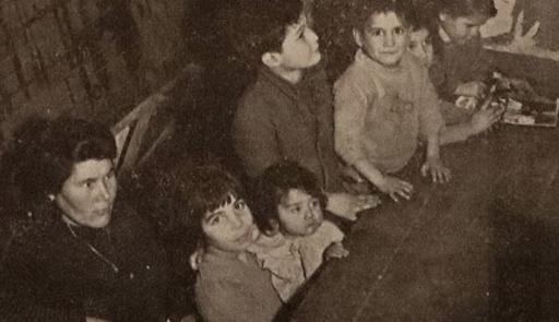 Foto in bianco e nero di una donna e numerosi bambini seduti attorno ad un tavolo