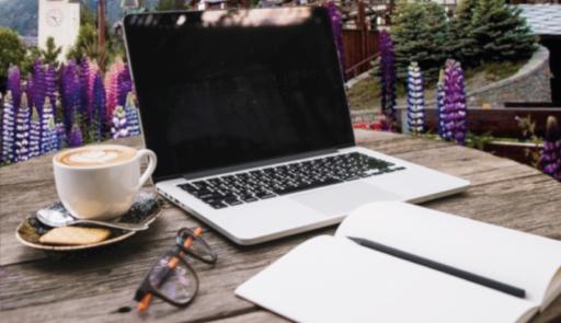 tavolo in legno con appoggiato computer, cappuccino, occhiali e quaderno