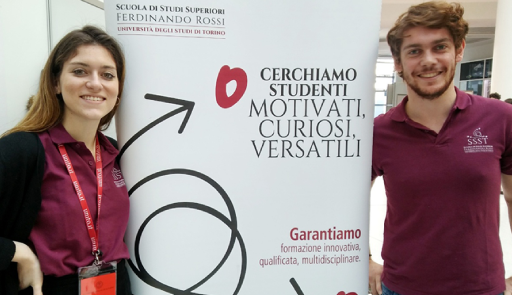 Una studentessa e uno studente a lato di un roll up che promuove la scuola di studi superiori