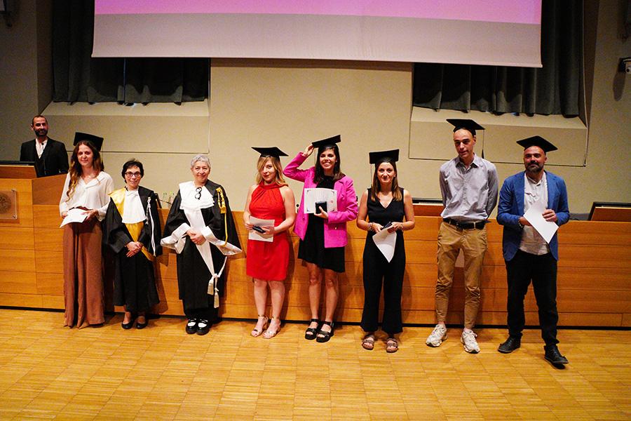Sei persone con indosso il cappello da laureato, la prorettrice e una professoressa in toga posano di fronte al fotografo nell'aula magna della Cavallerizza in occasione della Festa della Conoscenza - Migliori Laureate e Laureati
