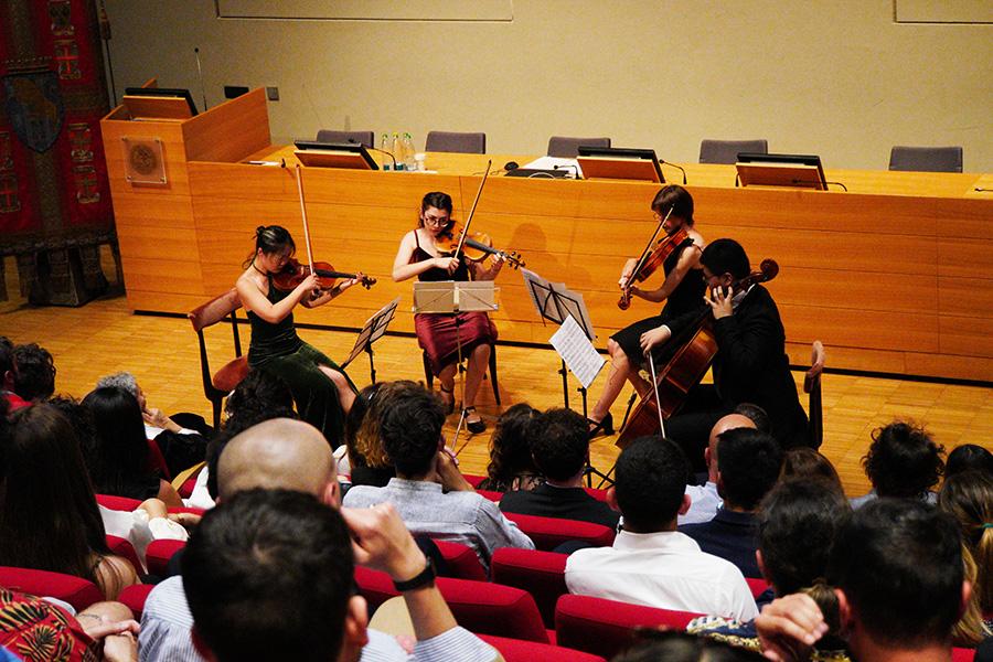 Quatto persone suonano il violino di fronte al pubblico della Festa della Conoscenza Migliori Laureate e Laureati nell'aula magna della Cavallerizza