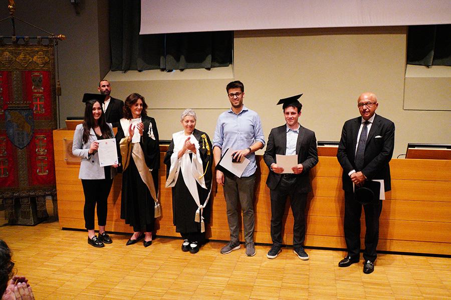 Tre persone con indosso il cappello da laureato, la prorettrice, una professoressa in toga e un professore posano di fronte al fotografo nell'aula magna della Cavallerizza in occasione della Festa della Conoscenza - Migliori Laureate e Laureati