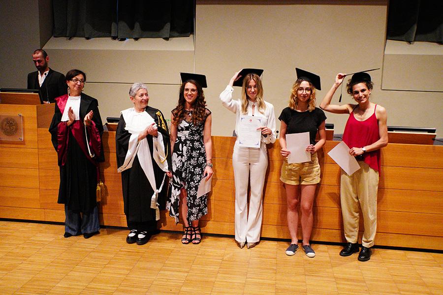 Quattro persone con indosso il cappello da laureato, la prorettrice e una professoressa in toga posano di fronte al fotografo nell'aula magna della Cavallerizza in occasione della Festa della Conoscenza - Migliori Laureate e Laureati