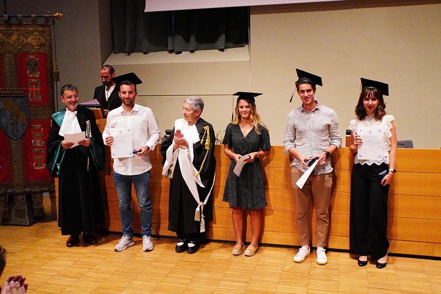 Quattro persone con indosso il cappello da laureato, la prorettrice e un professore in toga posano di fronte al fotografo nell'aula magna della Cavallerizza in occasione della Festa della Conoscenza - Migliori Laureate e Laureati