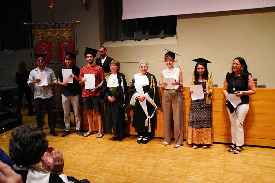 Sei persone con indosso il cappello da laureato, la prorettrice e una professoressa in toga posano di fronte al fotografo nell'aula magna della Cavallerizza in occasione della Festa della Conoscenza - Migliori Laureate e Laureati