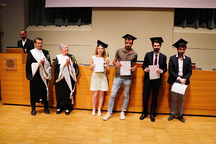Quattro persone con indosso il cappello da laureato, la prorettrice e un professore in toga posano di fronte al fotografo nell'aula magna della Cavallerizza in occasione della Festa della Conoscenza - Migliori Laureate e Laureati