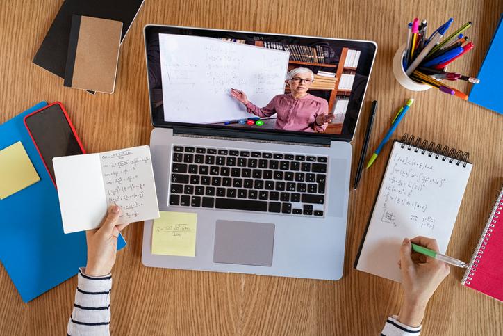 iStock / Getty Images Plus / Ridofranz Immagine con computer portatile sul tavolo accesso con videolezione a distanza. Intorno al computer sono poggiati telefono cellulare, block notes, libri e portabiro. Si intravvedono le mani di una studentessa che reggono sulla mano sinistra un block notes con appunti e sulla mano destra una biro che prende appunti su un altro block notes