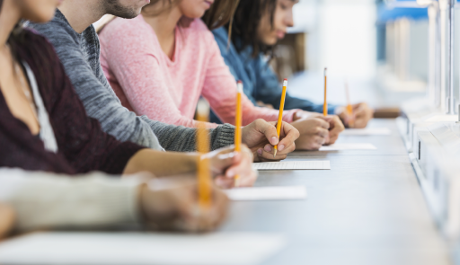 4 studenti in fila su una scrivania, con ciscuano che reggono in mano una matita intenti a scrivere su un foglio bianco