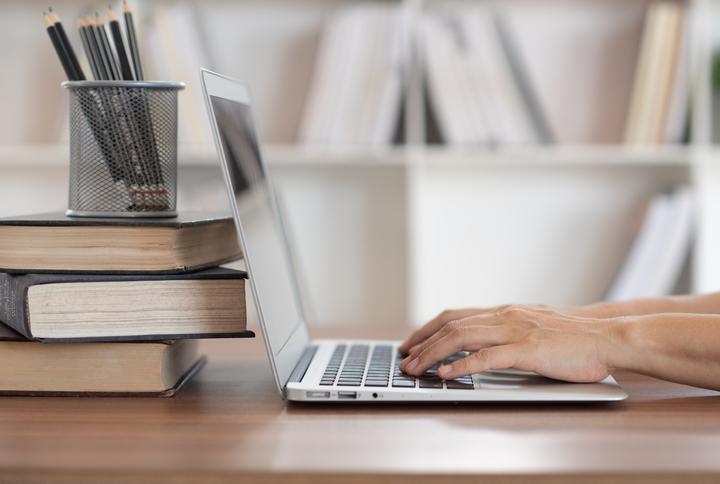 Immagine di profilo di un computer portatile con le mani di uno studente sulla tastiera e dietro il display pila di tre libri con sopra un portabiro