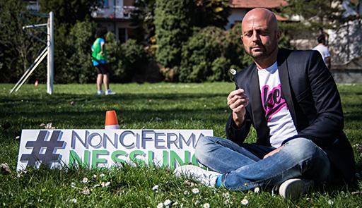 Foto di Luca Abete seduto su prato con un fiore in mano, di fianco è presente un cartello con la scritta:#NonCiFermaNessuno  