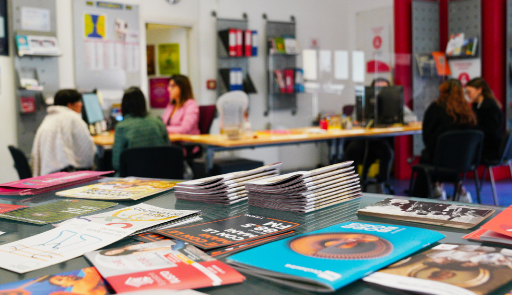 Immagine dei locali dell'infopoint con in primo piano diversi opuscoli su un tavolo e sullo sfondo alcuni studenti allo sportello