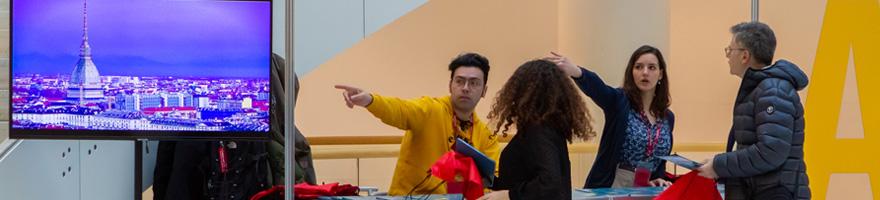 Due persone chiedono indicazioni a un desk informativo. Le due persone che danno informazioni indicano con la mano verso destra. A fianco del desk c'è un monitor con una visione panoramica di Torino
