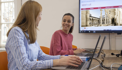 Due studentesse sedute ad una scrivania, una fianco all'altra, con sopra due portatili, che dialogano fra loro e con alle spalle un monitor che proietta la home page del portale di Ateneo