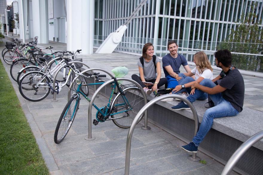Quattro ragazzi, di cui due femmine e due maschi, seduti una panchina a chiacchierare e accanto a loro le biciclette