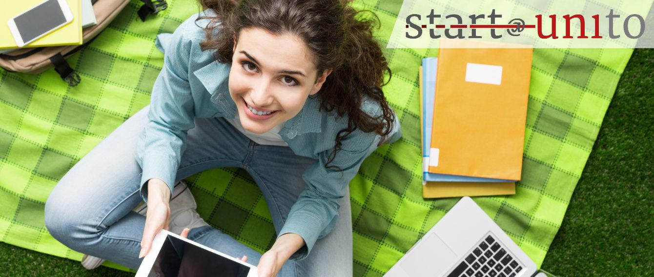 Ragazza sorridente, seduta su un prato con libri e pc portatile, logo start@unito